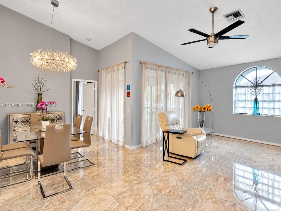 Open floor plan featuring vaulted ceilings, polished stone-finish tile flooring, a statement crystal chandelier, a sleek metal ceiling fan, and an arched window