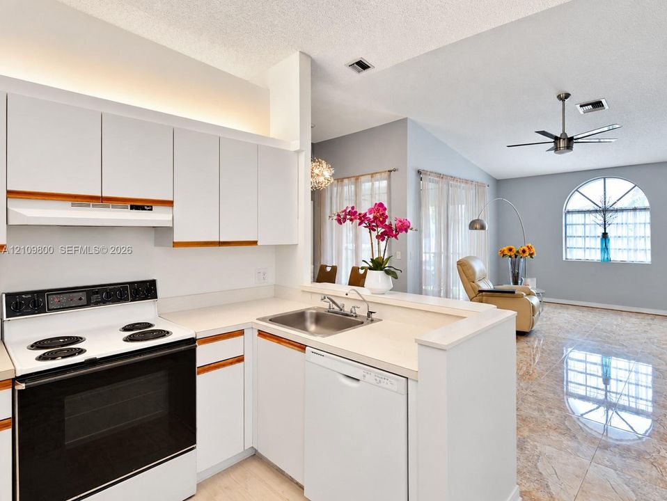 Kitchen featuring white cabinetry with wood trim, solid surface countertops, stainless steel sink with a goose-neck faucet, and white appliances including a range and dishwasher, complemented by large-format tile flooring and an arched window