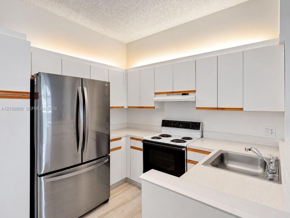 Kitchen featuring white flat-panel cabinetry with wood trim, stainless steel refrigerator, white electric range, integrated cove lighting, and a stainless steel sink with a high-arc faucet