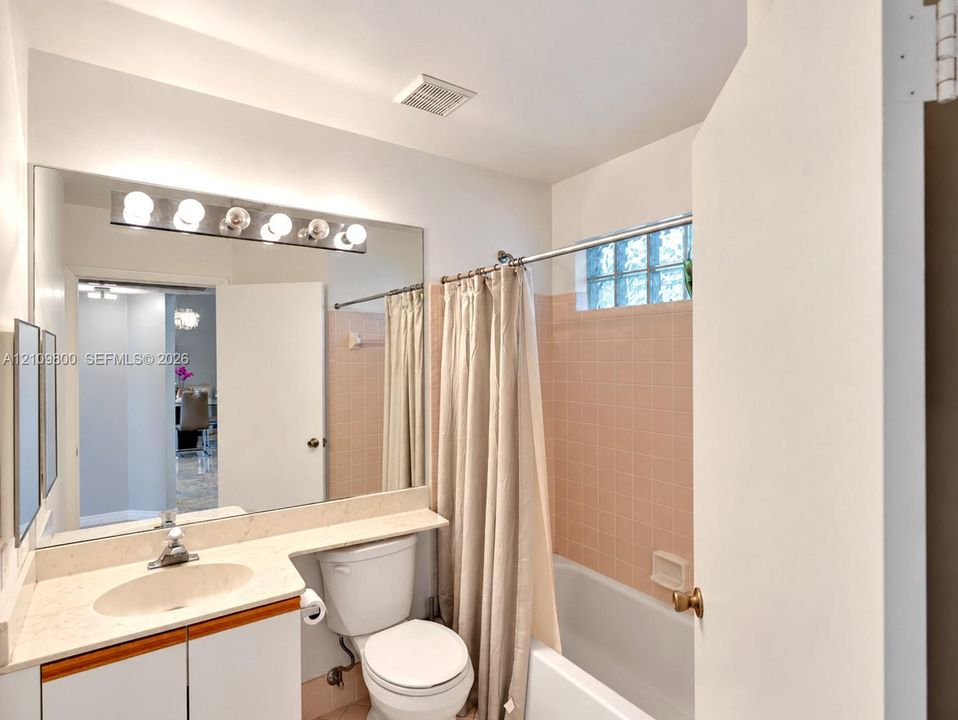 Bathroom featuring a stone-surface vanity with an integrated oval sink, a wall-spanning mirror with integrated multi-bulb lighting, and a bathtub surround finished with ceramic tile and glass block accent windows