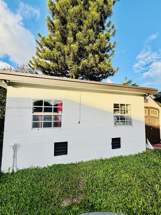 Exterior white masonry wall featuring two grid-pane windows, dark-toned shutters, and a rear storage structure with paneled doors