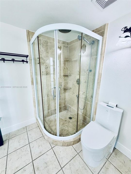Corner shower enclosure featuring a rounded glass door, integrated stone shelving, and overhead rainfall showerhead, complemented by a dual-flush toilet and large-format tile flooring