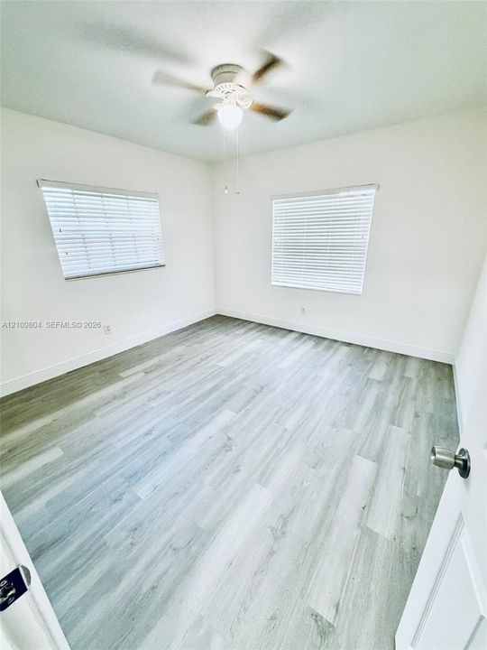 Neutral interior featuring wood-finish flooring, two dual-pane windows with horizontal blinds, and a ceiling-mounted fan with integrated lighting