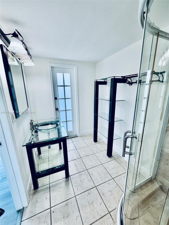 Glass-top vanity featuring a circular vessel sink, tiled flooring, glass-paneled exterior door, glass shelving unit with metal frame, and glass-enclosed shower with tiled surround