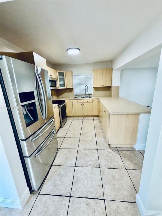 Kitchen featuring light wood cabinetry, stainless steel appliances, neutral solid-surface countertops, dual-basin stainless steel sink, and large-format tile flooring