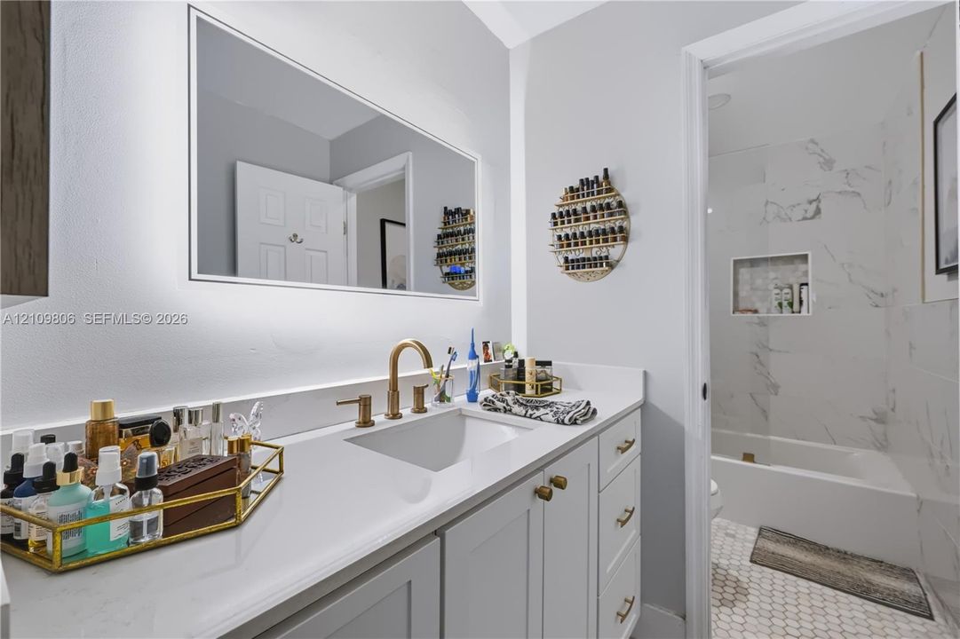 Bathroom featuring a vanity with quartz countertop, undermount sink, and brass-tone faucet hardware, complemented by an integrated backlit mirror and a tiled shower surround with a built-in recessed niche