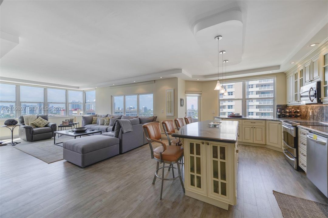 Open-concept kitchen featuring an expansive island with stone-surface countertop, custom cabinetry with glass-paneled detailing, stainless steel appliances, and wood-finish flooring throughout