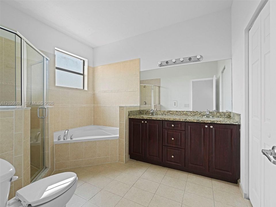 Bathroom featuring a dual-sink vanity with dark wood-finish cabinetry, stone-surface countertop, tile-surround corner soaking tub, separate glass-enclosed shower, and full-width vanity mirror