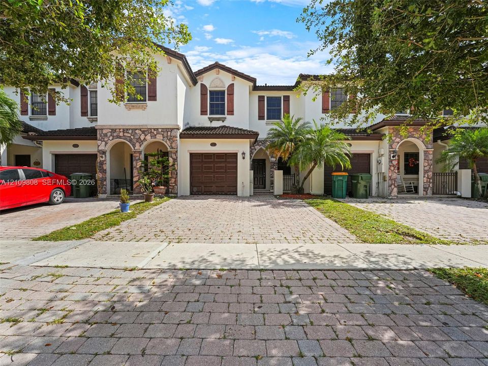 Multi-level townhouse exterior featuring light-toned stucco walls, decorative stone veneer detailing, arched entryway accents, brown-tone window shutters, and a paved driveway