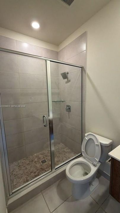 Walk-in shower with floor-to-ceiling tile surround, glass enclosure, integrated glass shelving, and pebble-textured shower floor, complemented by neutral tile flooring