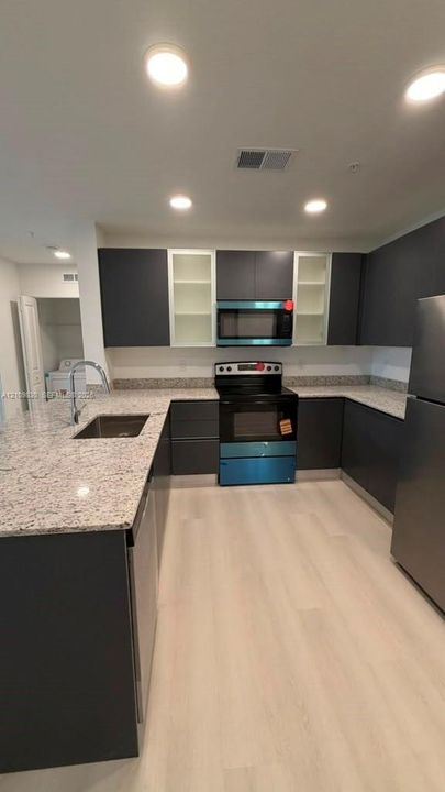 Kitchen featuring dark cabinetry with glass-front uppers, stone-patterned countertops, stainless steel appliances, a peninsula with an undermount sink, and light wood-finish flooring