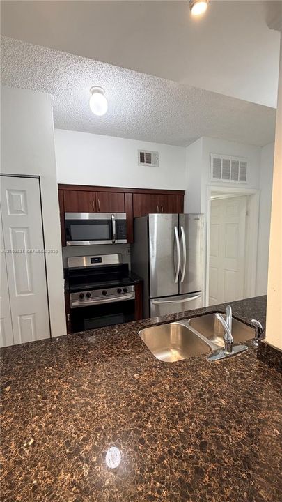 Kitchen featuring dark wood-finish cabinetry, stainless steel appliances, a multi-tone stone-finish countertop, and a double-basin stainless steel sink with a high-arc faucet
