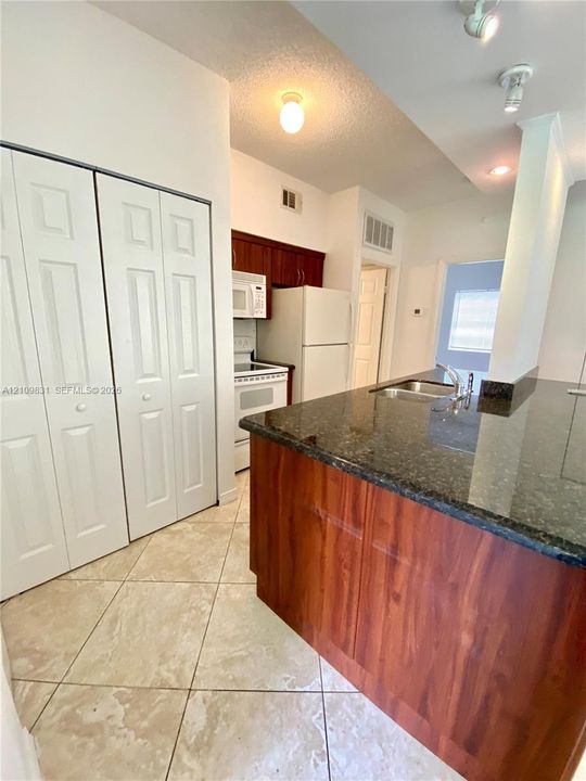 Kitchen featuring a dark stone-finish countertop, dual-basin stainless steel sink, wood-finish cabinetry, tile flooring, and white bi-fold closet doors