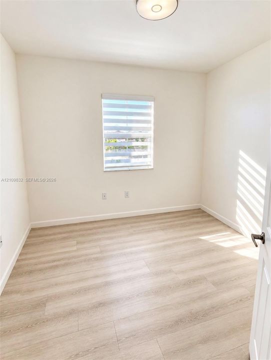 Neutral-toned interior featuring wood-finish flooring, a single window with horizontal blinds, and a flush-mount ceiling light fixture