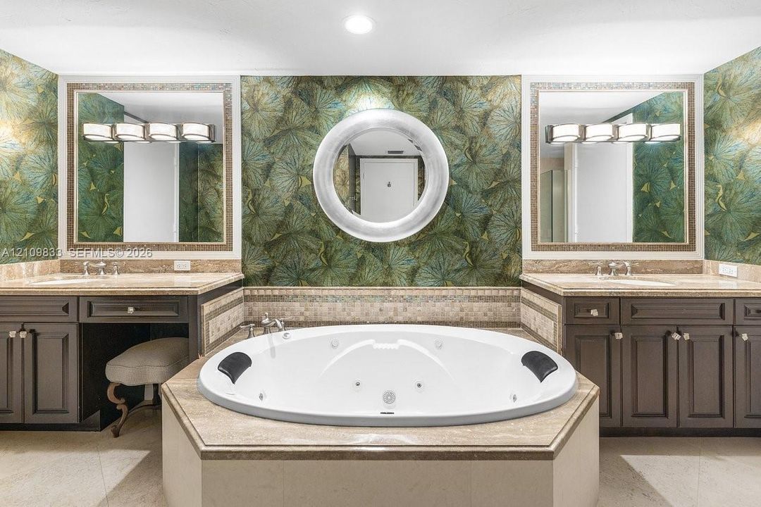 Dual vanities with stone countertops, jetted soaking tub, decorative wall covering, and two framed wall mirrors