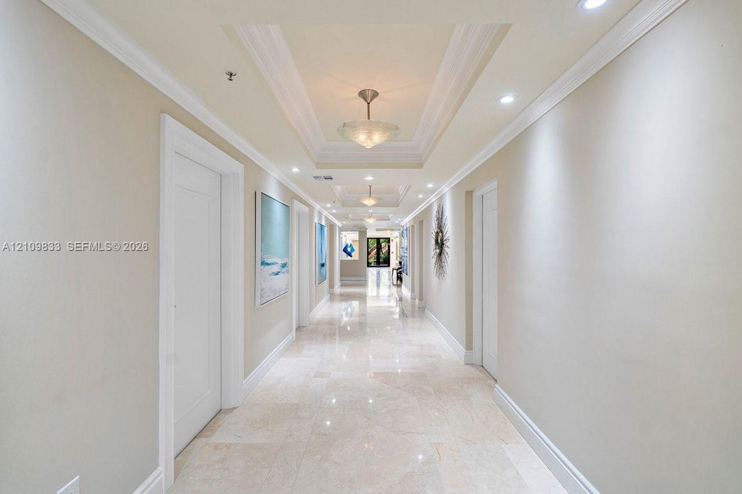 Grand gallery hallway featuring stone tile flooring, multi-tiered tray ceilings, recessed lighting, and decorative semi-flush mount fixtures