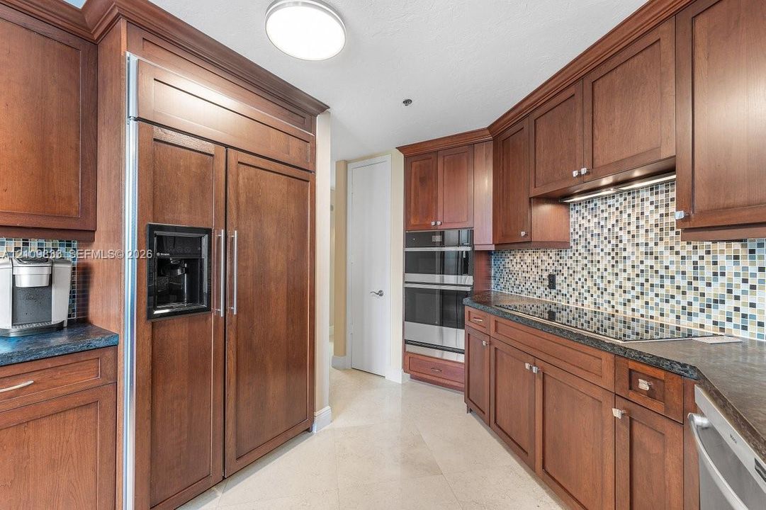 Kitchen featuring dark wood cabinetry, panel-front refrigerator, stainless steel built-in double ovens, multi-colored glass tile backsplash, and stone-finish countertops
