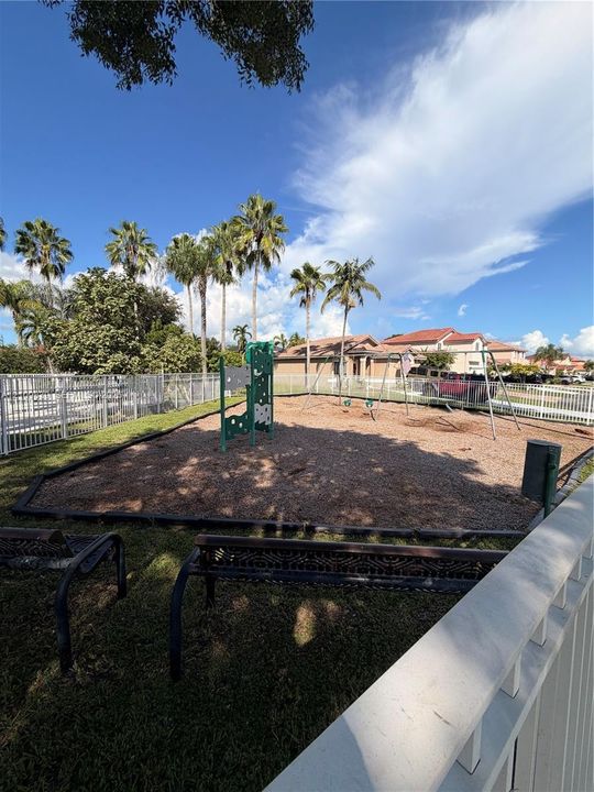Recreational area featuring a climbing structure, swing set, wood-chip ground cover, and perimeter fencing shaded by mature palm trees