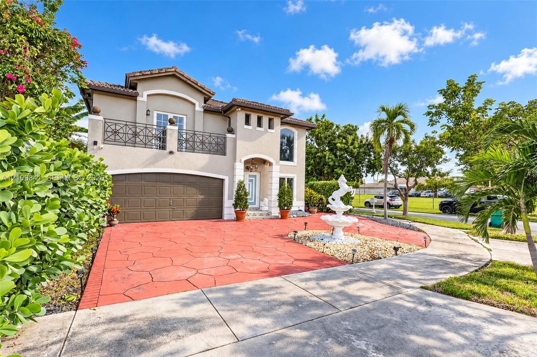 Two-story exterior featuring a stamped concrete driveway, decorative stone fountain, upper-level balcony with metal railing, and arched garage entry