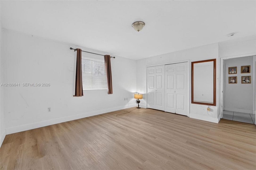 Room with wood-finish flooring, white walls, built-in double-door closet, wall-mounted wood-framed mirror, and window with blinds