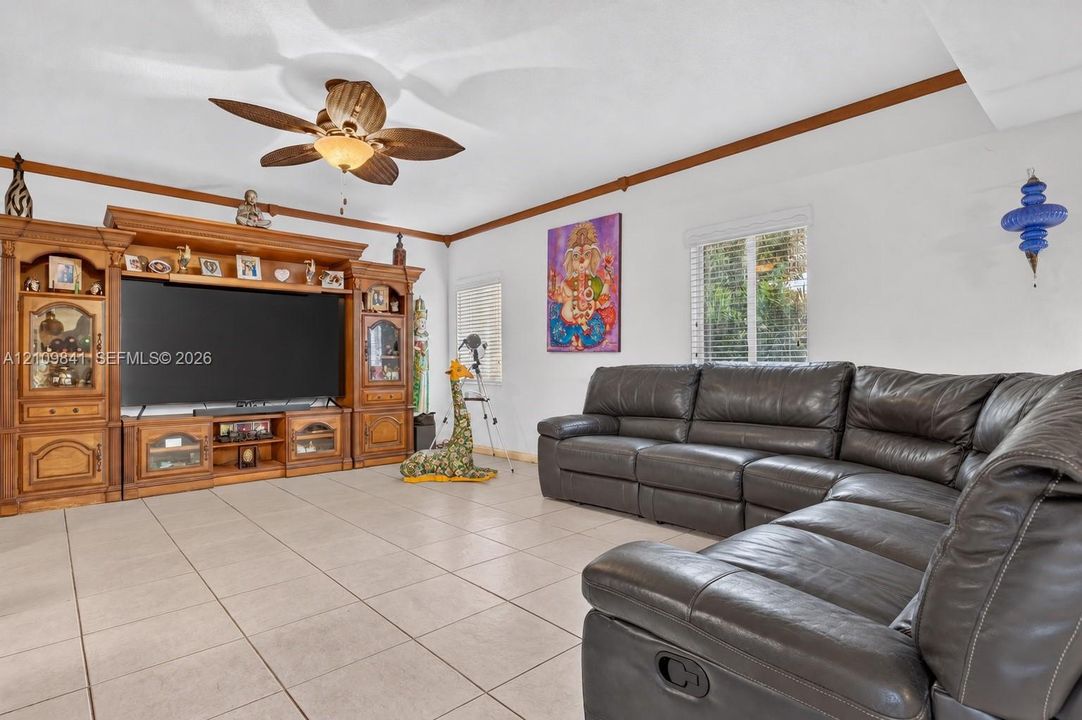 Living area featuring large-format tile flooring, crown molding, a multi-blade ceiling fan with integrated lighting, and an expansive built-in wood cabinetry unit
