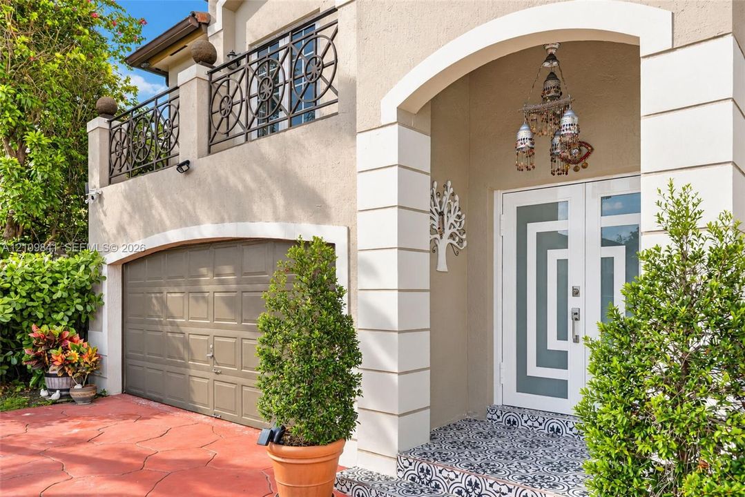 Exterior facade featuring a patterned tile entryway, double front doors with geometric glass inserts, a second-story balcony with decorative metal railings, and an arched garage entry