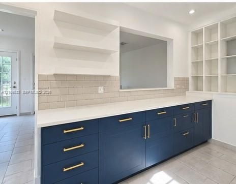 Navy blue cabinetry with gold-tone pulls, quartz countertops, subway tile backsplash, floating wall shelving, and built-in floor-to-ceiling cabinetry