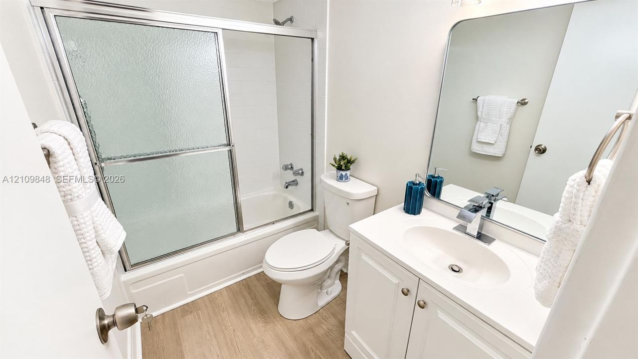 Bathroom featuring a white vanity cabinet with a solid-surface countertop, an integrated oval sink with a chrome single-lever faucet, a rectangular wall-mounted mirror, and a tub-shower combination with sliding glass doors and wood-finish flooring