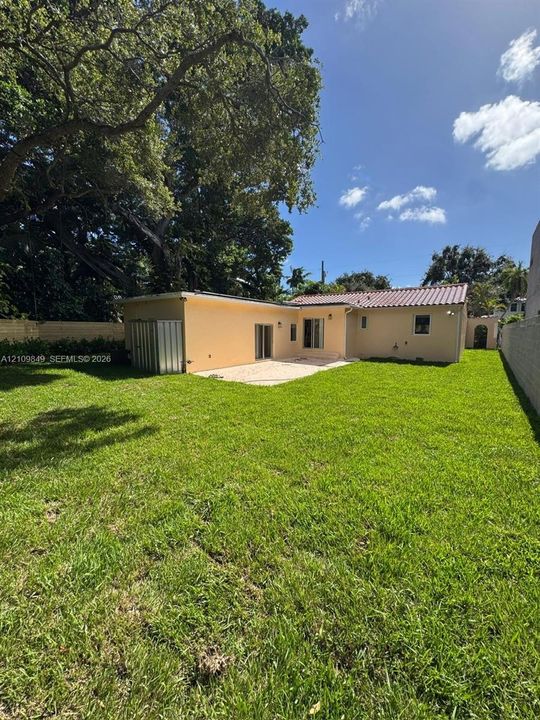 Expansive backyard featuring a manicured lawn, perimeter masonry walls, mature trees, a concrete patio, and a metallic storage structure