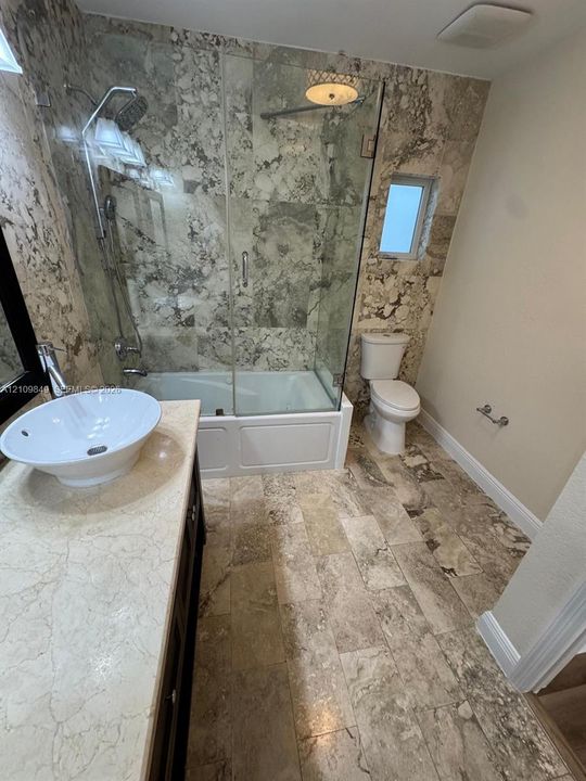 Bathroom featuring a vessel sink with a stone-top vanity, a glass-enclosed tub shower, and full-height stone-finish wall tile