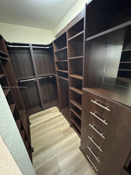 Custom walk-in closet featuring dark wood-finish cabinetry, extensive adjustable shelving, integrated hanging rods, a built-in drawer stack with brushed metal hardware, and wood-finish flooring