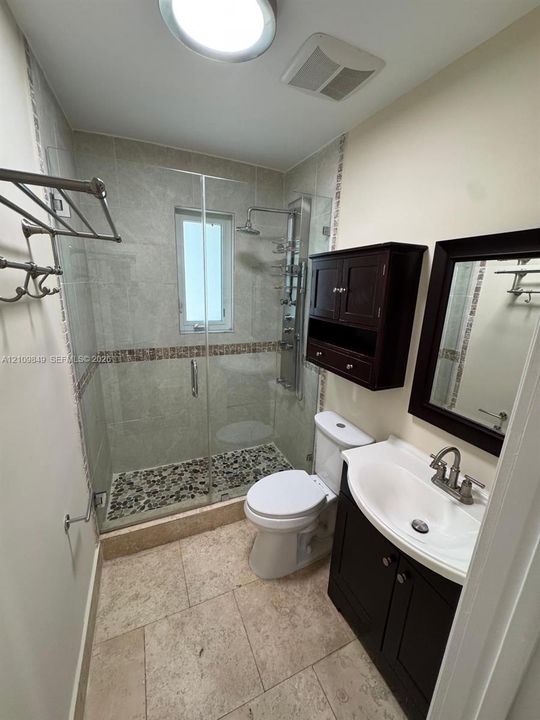 Bathroom featuring stone-tile flooring, a glass-enclosed shower with a pebble-stone floor, integrated shower panel system, dark wood-finish vanity with an oval basin, and matching wall-mounted cabinetry