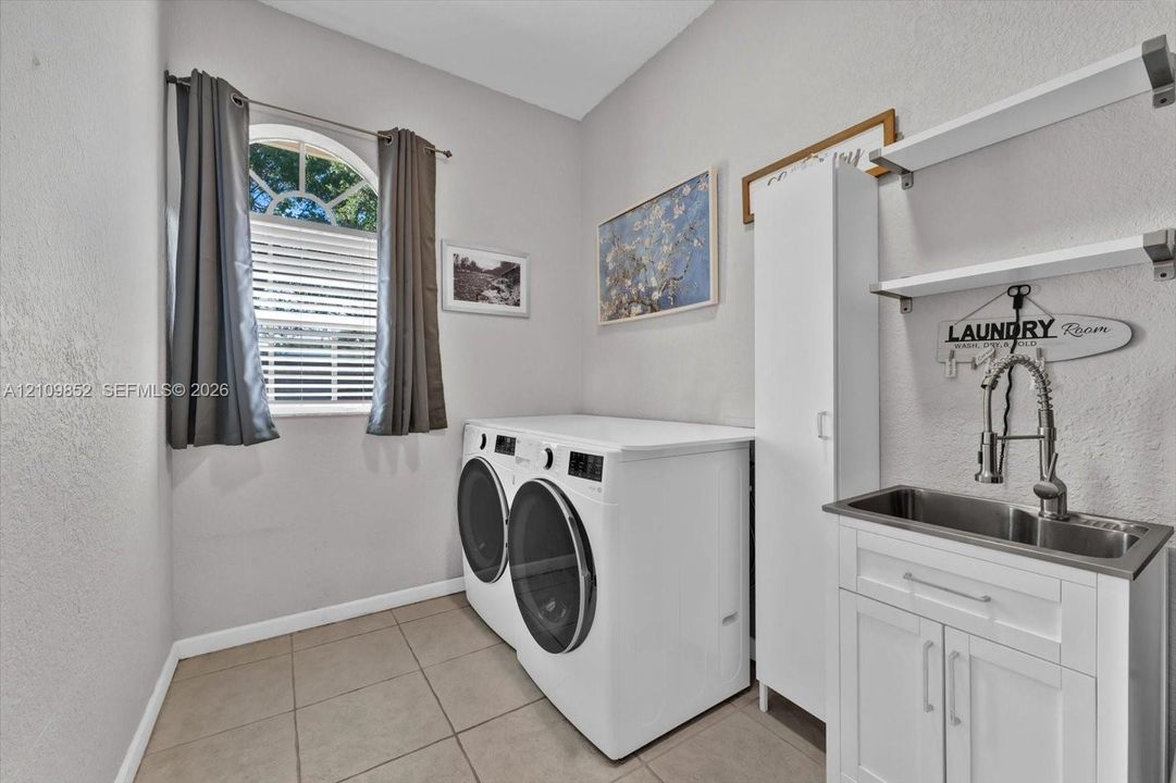 Laundry room featuring ceramic tile flooring, an arched window, a white utility cabinet, and a stainless steel sink with a high-arch pull-down faucet and wall-mounted shelving