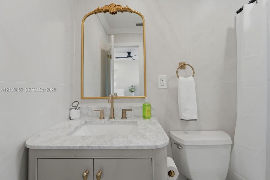 Bathroom featuring a light-toned vanity with stone-surfaced countertop, under-mount sink, brass-finish widespread faucet, arched decorative wall mirror, and porcelain wall tile