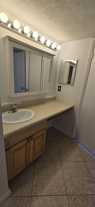 Bathroom vanity featuring light-finish wood cabinetry, a solid-surface countertop with an integrated basin, a tri-fold medicine cabinet, and a multi-bulb light fixture above large-format floor tile