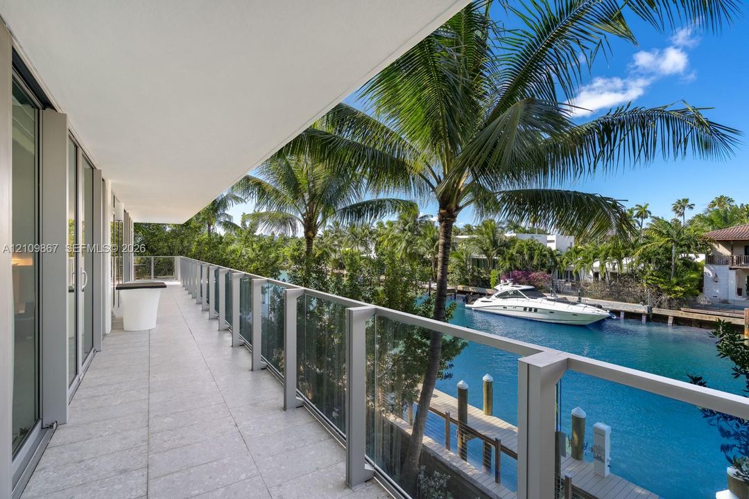 Covered balcony featuring stone-tiled flooring, glass panel railings, and metal framing, overlooking a canal with a private dock and mature palm trees