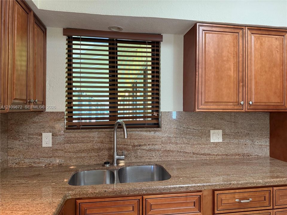 Kitchen featuring natural wood cabinetry, stone-surface countertop, matching stone backsplash, dual-basin stainless steel sink, and high-arc faucet