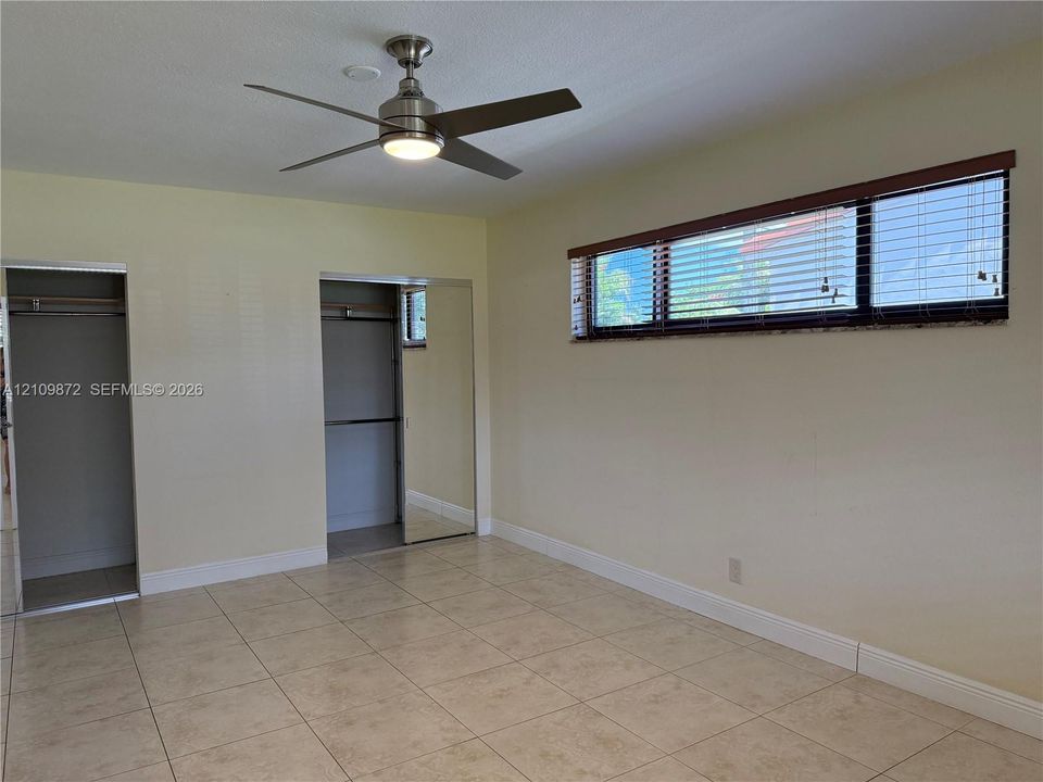 Neutral-toned tile flooring, brushed-metal ceiling fan with integrated light fixture, horizontal window with slat blinds, and dual closets with sliding mirrored doors