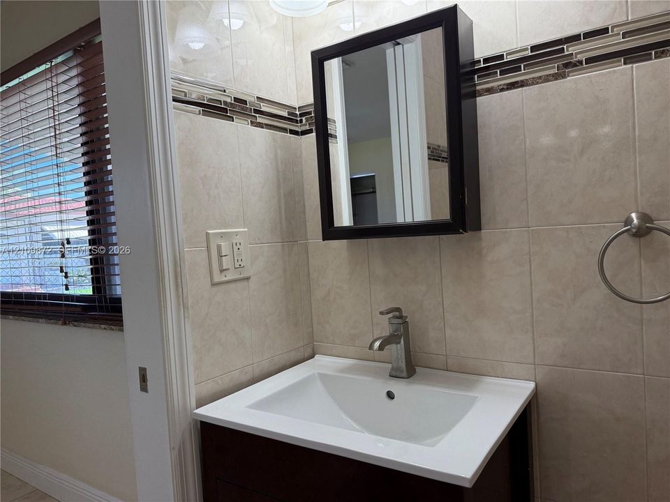 Dark wood-finish vanity with a white rectangular basin, metallic single-handle faucet, integrated medicine cabinet, and ceramic wall tiling with decorative glass tile inlay