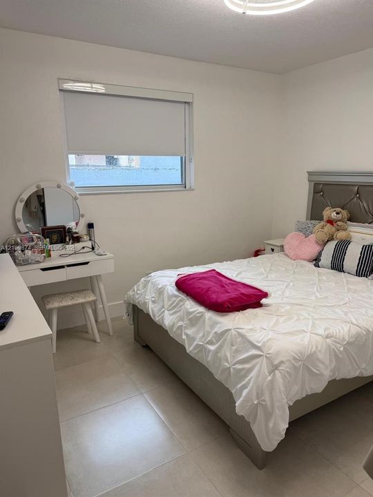 Bedroom featuring large-format ceramic tile flooring, a window with a retractable roller shade, and a ceiling-mounted light fixture