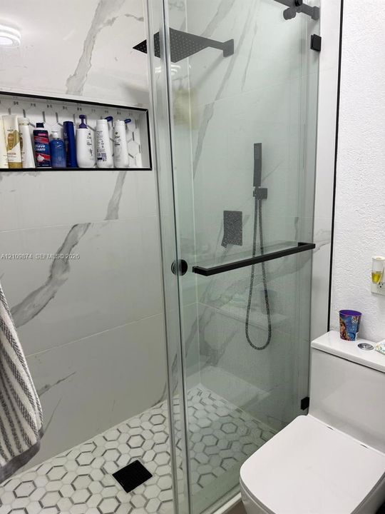 Walk-in shower featuring large-format wall tile with a veined pattern, hexagonal mosaic floor tile, glass enclosure with matte black hardware, integrated storage niche, and a matte black rainfall showerhead