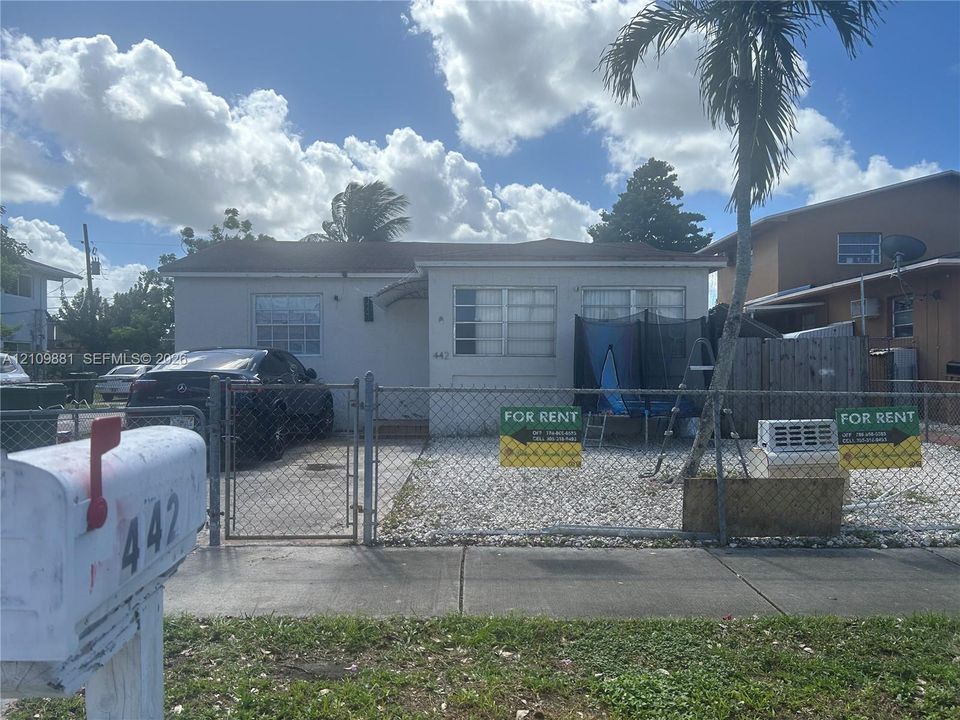 Single-story exterior with a concrete facade, horizontal grid windows, paved parking area, and a chain-link perimeter fence