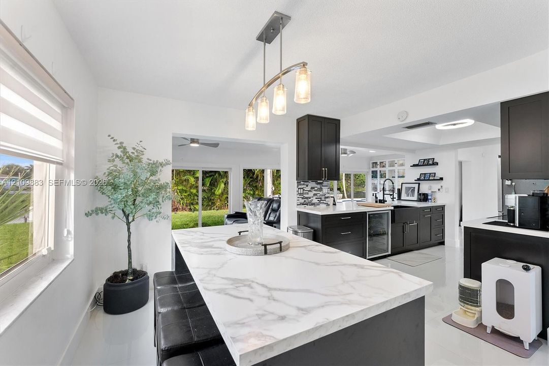 Kitchen featuring an expansive island with a light-toned stone-finish countertop, dark cabinetry, an integrated beverage refrigerator, a farmhouse-style sink, and a multi-light pendant fixture