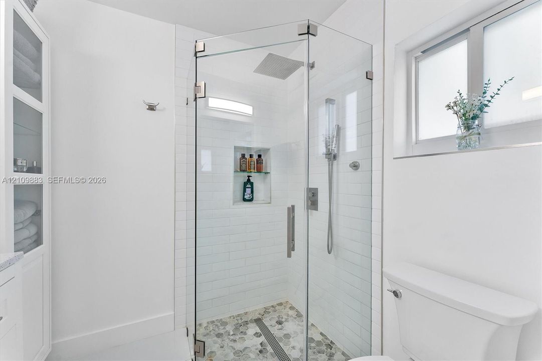 Walk-in glass shower with subway tile surround, integrated shelving, rainfall shower head, and stone-finish mosaic flooring