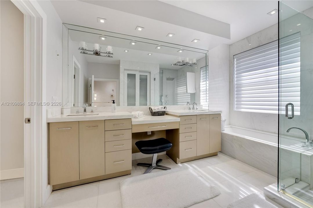 Dual vanity with integrated stone-finish countertop, recessed lighting, expansive wall-to-wall mirror, glass-enclosed shower, and a built-in soaking tub with stone-finish surround