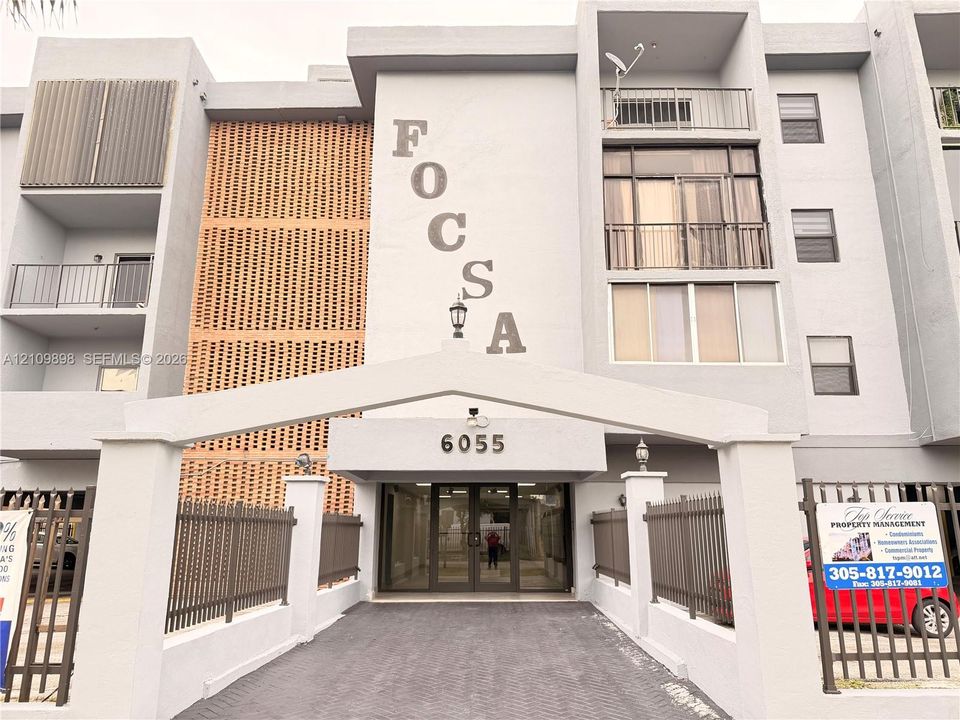 Multi-story residential building exterior featuring a structured gated entry, paver driveway, decorative brick-screen facade elements, and private balconies with metal railings