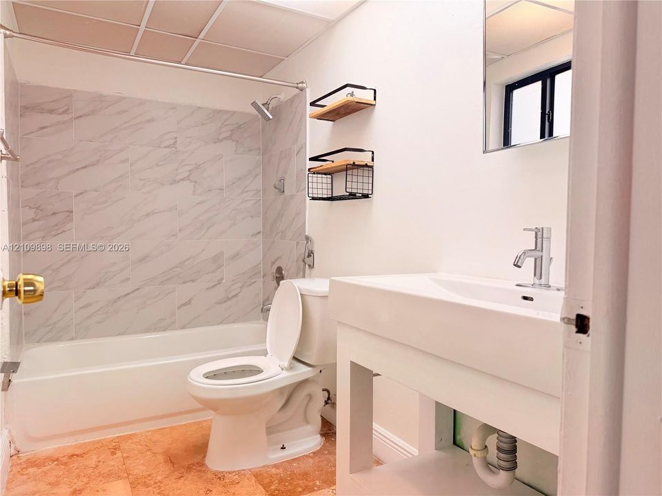 Bathtub shower combination with stone-patterned tile surround, white pedestal-style vanity with single-handle faucet, and tile flooring
