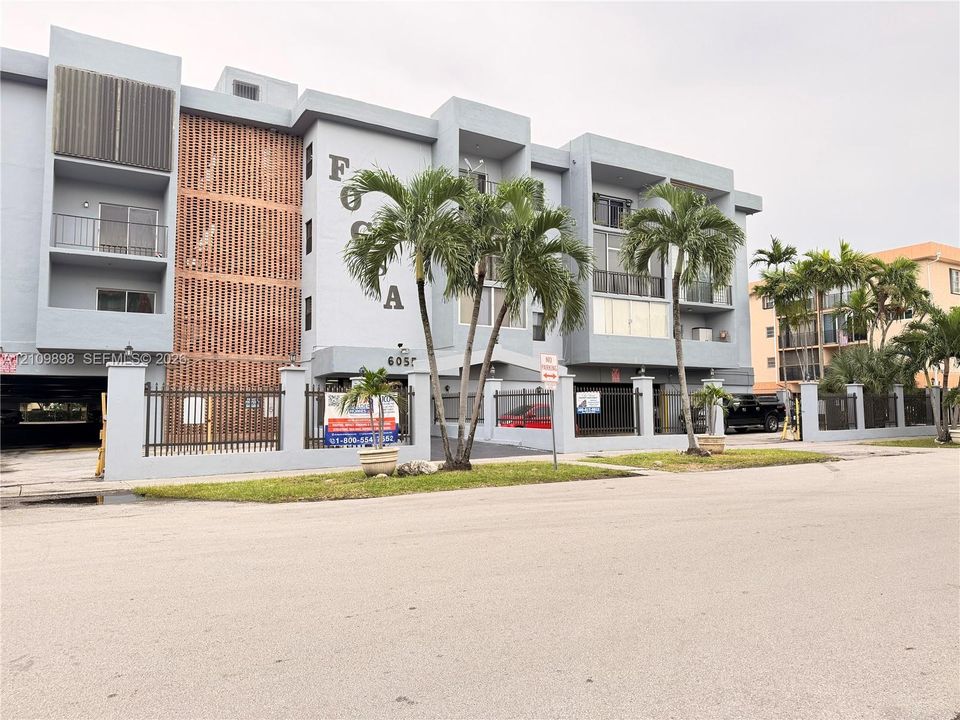 Multi-story building exterior featuring concrete construction, decorative brick accent panels, private balconies, gated ground-level parking, and mature palm landscaping