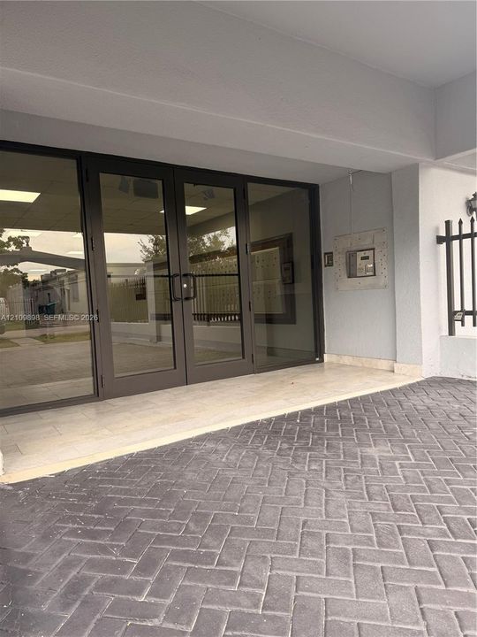 Covered entrance featuring double glass doors, a stone-tiled threshold, and a herringbone-patterned paved driveway with a wall-mounted intercom system