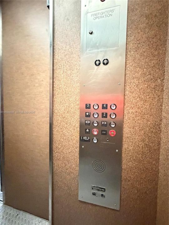 Elevator interior featuring a brushed metal control panel with integrated push-button operation and emergency communication system, set against textured, neutral-toned wall surfacing